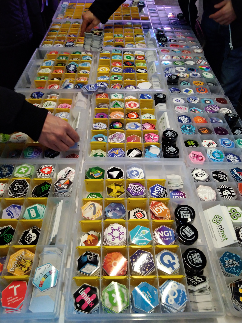 a table entirely laden with boxes containing hexagonal stickers neatly laid out. Some hands are visible rifling through the assorted stickers.